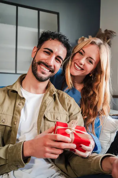 Vertical. Real young couple smiling looking at camera showing a birthday gift. Man holding a valentines surprise present with his wife. Boyfriend and girlfriend celebrating the relation anniversary