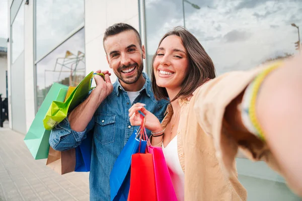 Neşeli genç beyaz bir çift, gülümsüyor ve alışveriş merkezinde alışveriş torbaları tutarak selfie çekerken eğleniyor. Müşteriler mutlu bir ifadeyle öne bakıyor. Fotoğraf çeken arkadaşlar.