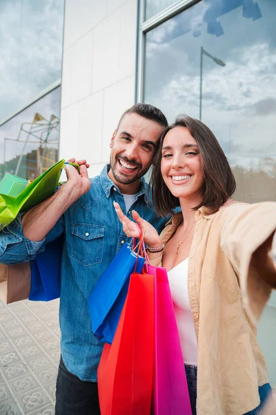 Vertical. Young caucasian real couple smiling and having fun taking a selfie portrait holding shopping bags at city store mall. Customers staring front with happy expression. Friends shooting a photo