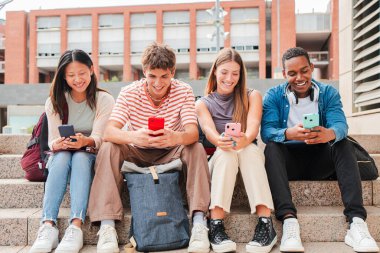 Bir grup çok ırklı lise öğrencisi üniversite kampüsündeki bir akıllı telefondan sosyal medya uygulamasını izlerken eğleniyor. Genç sınıf arkadaşları cep telefonu kullanıyor. Mutlu arkadaşlar internete telefonla bakıyorlar.