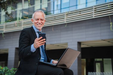 Takım elbise ve kravat takmış, gülümseyen beyaz yakalı bir işçi, dizüstü bilgisayar ve cep telefonuyla bir binanın ön cephesinde oturuyor. İnternette çalışmak için cep telefonu ve bilgisayar kullanan mutlu bir iş adamı. Yüksek