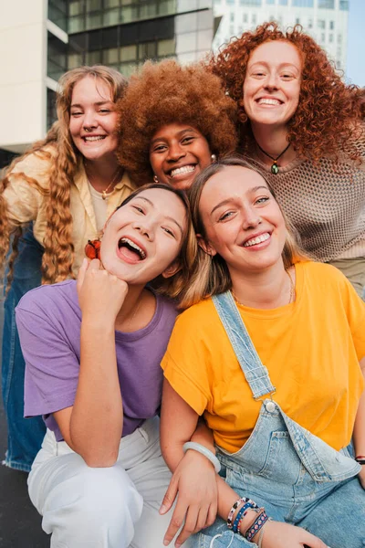 Vertical social group of real young women are smiling and posing in front of a building, showing happy facial expressions, having fun while traveling as a community, enjoying leisure time together