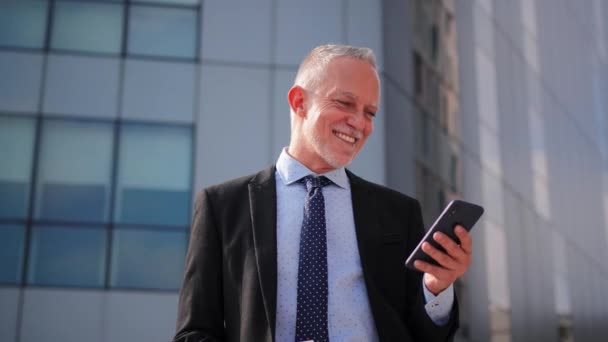 一位穿着西装 领带和连衣裙的成熟商人站在一幢大楼前 手持手机时面带微笑 公司高级首席执行官在工作场所使用智能手机 — 图库视频影像