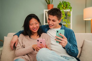 Oturma odasındaki koltukta oturan uygulamaları aramak için cep telefonu kullanan çok ırklı evli bir çift. Genç insanlar gülümsüyor ve akıllı bir telefonla birlikte eğleniyorlar. Sosyal medyada iki arkadaş paylaşıyor