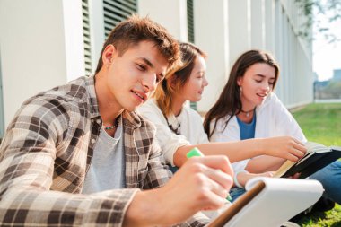 Genç öğrenci not defterine kalemle notlar yazıyor. Üniversite kampüsünün bahçesinde sınıf arkadaşlarıyla oturuyor. Liseli çocuk sınav için ders okuyor ve çalışıyor. Takım çalışması arkadaşlar