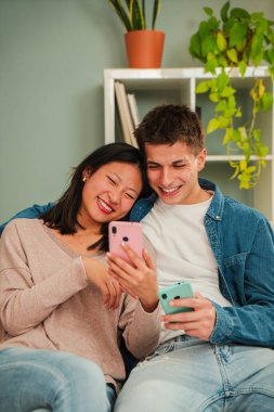 Dikey. Çok ırklı evli çift oturma odasındaki koltukta oturan uygulamaları aramak için cep telefonu kullanıyor. Genç insanlar gülümsüyor ve akıllı bir telefonla birlikte eğleniyorlar. Sosyal medyada arkadaşlar paylaşıyor