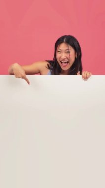 Dikey afiş. Asyalı genç bir kadın boş bir tahtayı kırmızı zemine doğrultuyor. Tavsiye fotokopi alanı modeli. Çinli genç kız pembe arka plan duvarında aşağıdaki ilan panosunu gösteriyor.