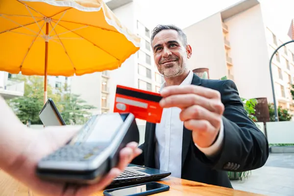Happy mature business man paying bill using a contactless credit card in a restaurant. Handsome mid adult male customer smiling holding a creditcard and giving a payment transaction to the cashier