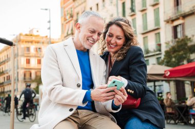 Orta yaşlı bir çift oteli cep telefonuyla arıyor. Dışarıda oturan bir uygulama kullanıyor. Olgun insanlar sosyal medya uygulamasında cep telefonuyla eğleniyorlar. Akıllı telefonlu orta yaşlı arkadaşlar.