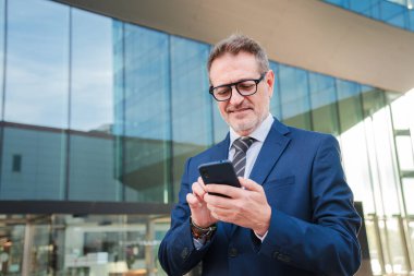 İş yerinde akıllı telefon kullanarak gülümseyen yakışıklı bir iş adamı. Gerçekten başarılı bir yönetici erkek ya da CEO cep telefonundaki bir finans uygulamasını izliyor. İş adamı elinde gülümseyerek bir cep telefonu tutuyor