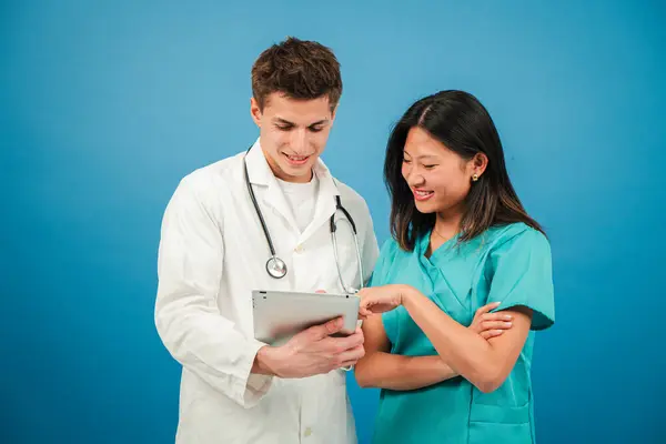 Two young adult doctors using a digital tablet to watch a diagnosis at blue background wall. Surgeon and cardiologist working with a touchpad. Specialist team. Nurse and assistant doc or physician