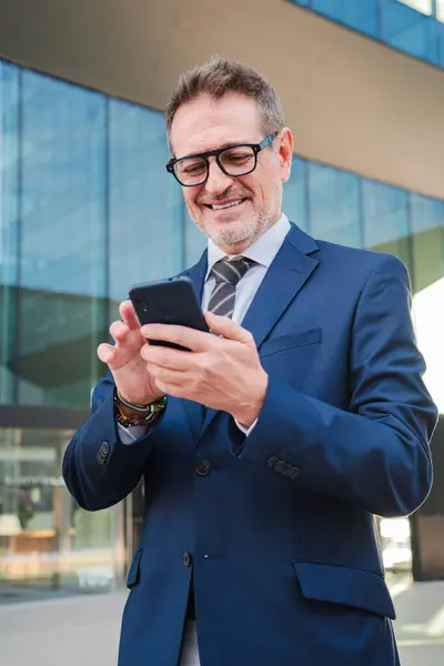 Vertical. Corporate business man smiling using a smartphone at workplace. Real successful executive male or ceo watching a finance app on his cellphone. Businessperson holding a mobile phone smiling