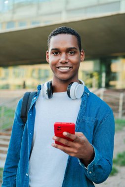 Kameraya bakıp elinde akıllı telefon tutan Afrikalı bir Amerikalının dikey portresi. Gerçek bir lise öğrencisi cep telefonu kullanıyor ve üniversite kampüsüne dik dik bakıyor. Telefonu olan adam.