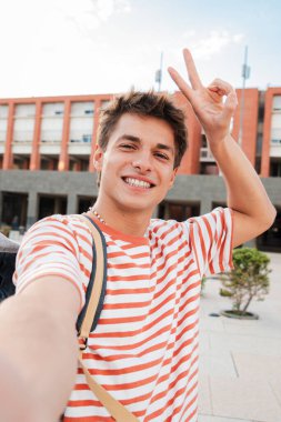 Kampüste kameraya bakıp gülen beyaz bir gencin dikey selfie portresi. Gerçekten yakışıklı bir genç adam gülüyor ve önüne bakıyor. Olumlu ifadeli neşeli erkek