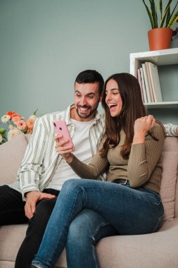 Dikey. Oturma odasındaki kanepede cep telefonuyla internette gezinen bir çift. İnternette cep telefonu uygulaması ile bahis oynayan iki arkadaş. Erkek arkadaş ve kız arkadaş ev arıyor ya da rezervasyon yaptırıyor