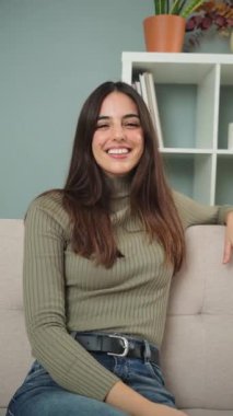 Dikey görüntü. Beyaz dişleriyle kameraya bakıp eğlenen mutlu beyaz kız öğrenci. Neşeli ve sevimli genç esmer kadın evinde bir kanepede oturmuş gülüyor.