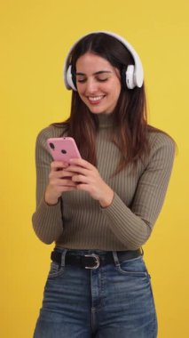 Dikey. Gülümseyen mutlu genç kadın akıllı telefonunu kullanıyor ve internette sohbet ediyor ya da sarı arka planda internette geziniyor. Genç beyaz kadın cep telefonu uygulamalı komik videolar izliyor.