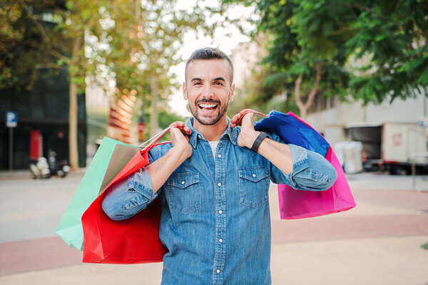 Only one young adult guy carrying shopping bags after purchase clothes on a mall store. Real happy male customer showing the packages that he buy on black friday with discount. Shopaholic man smiling