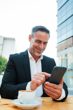 Dışarıda cep telefonunu kullanan bir iş adamının dikey portresi. Olgun erkek akıllı bir telefon cihazıyla internette geziniyor. Orta yaşlı girişimci gülümseyip cep telefonuyla mesaj atıyor. Yüksek