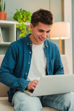 Dikey. Kanepede dizüstü bilgisayarla çalışan mutlu bir serbest çalışan. Oturma odasında bilgisayarla gülümseyen ve internette gezinen genç bir adam. Girişimci banka hesaplarına ve yatırımlarına bakıyor. Yüksek