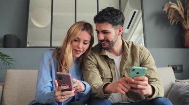 Young female holding a credit card, shopping online and her husband pointing her smart phone. Adult couple searching a sale on a cellphone social media app sitting on a couch at home living room. High