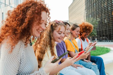 Çeşitli gençlerden oluşan canlı bir grup açık havada, akıllı telefonlarıyla meşgul bir şekilde birlikte oturuyorlar. Hepsi gülümsüyor ve zamanlarının tadını çıkarıyorlar. Modern gençlerin dijital bağlantısını sembolize ediyorlar.
