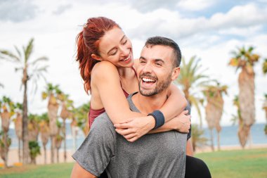 Man smiling and carrying his girlfriend piggyback after their cardio workout. Young athletic couple enjoying together, hugging and looking at each other affectionately. Healthy runner people. High clipart