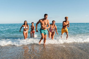 Sahilde koşuşturan bir grup çok ırklı arkadaş. Mutlu gençler sudan hızla çıkıp deniz kıyısına kadar yarışarak eğleniyorlar. Gençler mayo ile denizde oynuyorlar.