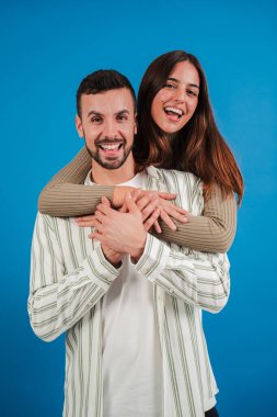 Dikey genç çift gülüyor ve mavi stüdyo arka planına bakıyor. Gerçek kız arkadaşı erkek arkadaşına sarılıyor ve birlikte gülümsüyor. Mutlu kadın yakışıklı ortağını kucaklıyor.