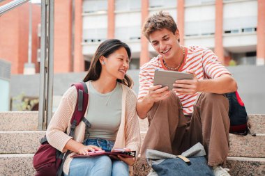 Üniversite kampüsünün merdivenlerinde oturan iki genç ders çalışıyor ve tablet kullanarak ödev yapıyor. Sınıf arkadaşları akademik bir proje için fikir paylaşıyorlar, dokunmatik bir dokunmatik altlıkla çalışıyorlar. Üniversite öğrencileri