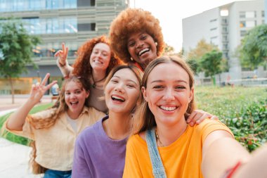 Sosyal medyada paylaşmak için akıllı telefonuyla selfie çeken bir grup çok ırklı genç kadın. Gerçek genç kızlar hafta sonları eğlenirken hatıra fotoğrafı çektirip gülümsüyorlar.