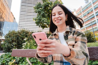 Genç İspanyol kadın cep telefonuyla mesaj atıyor ve mesaj yolluyor. Latin kız dışarıda internette gezinmek için akıllı telefon kullanarak eğleniyor. Genç bir kadın sosyal medyanın uygulamasında sohbet ediyor.