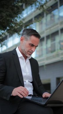 Finansal bölgenin dışında dizüstü bilgisayarla çalışan yetişkin bir iş adamının dikey portresi. Açık havada iş e-postalarına cevap vermek için not defteri kullanan yakışıklı orta yaşlı girişimci