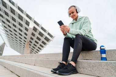 Olgun bir sporcu kadın kulaklıkla müzik dinlerken gülümsüyor ve egzersiz uygulamasını izlemek için akıllı telefonunu kullanıyor. Kardiyo çalışmasından sonra dinleniyor. Atletik kadın antrenmandan sonra gevşedi. Yüksek