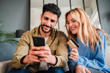 Genç bir çift cep telefonundaki bir teslimat uygulamasını kullanarak internetten yemek sipariş ediyor. Kadın ve erkek evlerindeki kanepede oturuyor, bir kredi kartı ve bir cep telefonuyla seyahat için internet üzerinden ödeme yapıyorlar. Yüksek