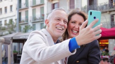 Evli, olgun bir çift, gülümsüyor ve tatilde telefonla selfie çekerken eğleniyor. Orta yaşlı bir adam ve kadın gezerken ve cep telefonuyla fotoğraf çekerken eğleniyorlar.