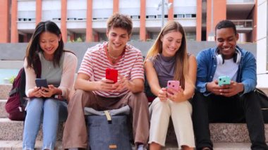 Sınav notlarını kontrol etmek için cep telefonu kullanan bir grup çok ırklı genç öğrenci. Genç sınıf arkadaşları üniversite kampüsünün merdivenlerinde otururken cep telefonu uygulaması ile internette geziniyorlar.