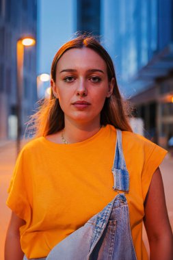 Geceleri dışarıda dikilen, ciddi, beyaz bir kadının dikey portresi. Düşünceli, gündelik kıyafetler giyen genç bir kadın sokakta kendine güvenen bir ifadeyle kameraya bakıyor. Yüksek