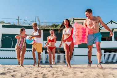 Bir grup arkadaş, hepsi mayo giyiyor, yaz tatili boyunca sahilde neşeyle koşuyorlar. Çok ırklı turistler kıyıya doğru koşan deniz tatillerinin tadını çıkarıyorlar. Yüksek