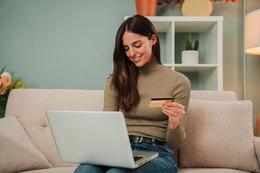 Genç beyaz bir kadın evdeki kanepede oturan kredi kartıyla online alışveriş yapmak için dizüstü bilgisayar kullanıyor. Kadın oturma odasındaki kanepede bilgisayarlı bir teslimat arıyor. İnternetten kız alışverişi. Yüksek