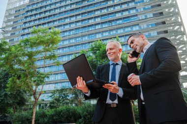 Dışarıda dizüstü bilgisayarla çalışan iki iş adamı, şehir merkezindeki finans bölgesinde duruyorlar. Dışarıdaki bir bilgisayarla daktilo eden ve e-posta gönderen iş adamları. Takım elbiseli 2 yönetici erkek.