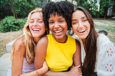 Gülümseyen ve kameraya bakan bir grup genç kadın. Üç çok ırklı genç kız birlikte eğleniyor. Neşeli kadınlar önlerine bakıp gülüyorlar. Kadınsı kadınlar tatillerinin tadını çıkarıyorlar.