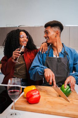 Dikey. Afro-Amerikalı çift, bir bardak kırmızı şarabın tadını çıkarırken evlerinin mutfağında vegan yemekleri pişiriyor. İki genç evde sağlıklı bir yemek hazırlamak için sebze kesiyor. Yüksek