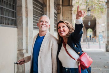 Olgun bir çift, bir bavulla geziyor ve işaret ediyorlar, tatillerinin tadını çıkarıyorlar. Orta yaşlı çift Avrupa şehir merkezini gezerken eğleniyor. Yetişkinler tatil yolculuklarının tadını çıkarıyorlar. Yüksek