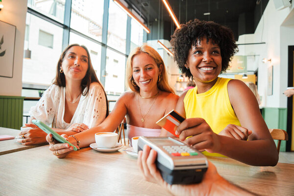 A cheerful young African American woman is settling her bill with a contactless creditcard at a restaurant. She smiles as she hands over her credit card to complete the payment with the cashier. High