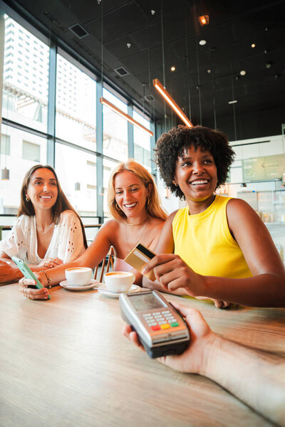 Vertical. Young African American woman is settling her bill with a contactless creditcard at a restaurant. She smiles as she hands over her credit card to complete the payment with the cashier. High