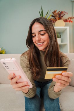 Dikey genç bir kadın akıllı bir telefonla ev koltuğunda oturan kredi kartıyla online alışveriş yapıyor. Kadın oturma odasındaki kanepede cep telefonuyla teslimat bekliyor. İnternetten alışveriş yapan kızlar
