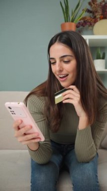 Dikey. Genç bir kadın evdeki koltukta oturan bir kredi kartıyla online alışveriş yapmak için akıllı bir telefon kullanıyor. Kadın oturma odasındaki kanepede cep telefonuyla teslimat bekliyor. İnternetten alışveriş yapan kızlar