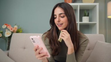 Kafkasyalı genç bir kadın evdeki koltukta oturan kredi kartıyla online alışveriş yapmak için akıllı telefon kullanıyor. Kadın oturma odasındaki kanepede cep telefonuyla teslimat bekliyor. İnternetten alışveriş yapan kızlar