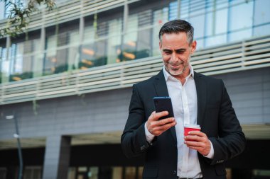 Deneyimli bir İşadamı: iş ve rahatlama arasında bir denge anı yakalamak için ofis dışında kahve içerken cep telefonunu kontrol eden olgun bir profesyonelin portresi. Yüksek